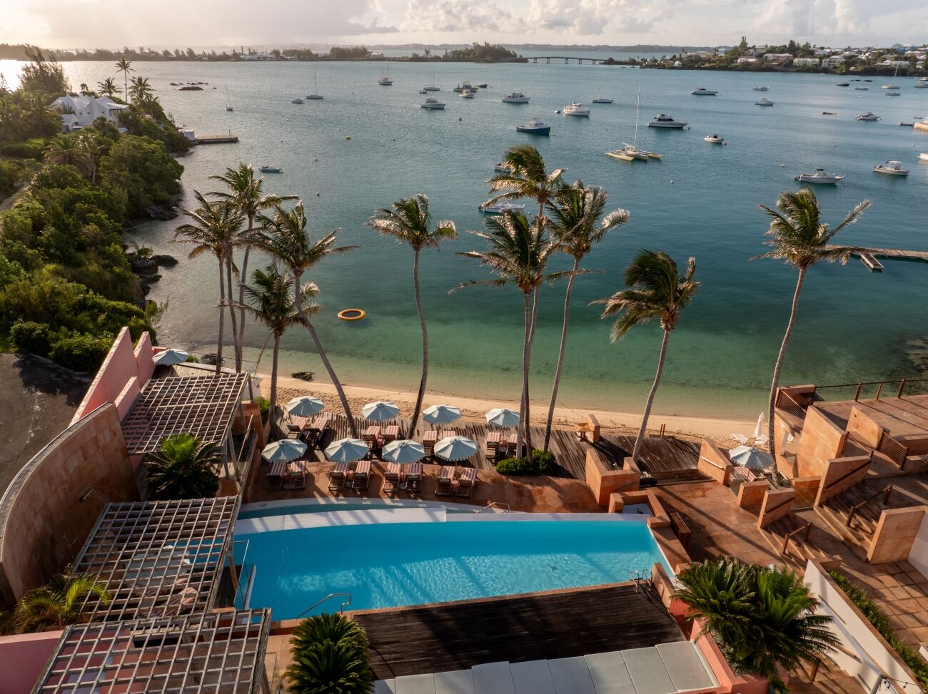 Colorful coastal resort with a pool, palm trees, beach, and boats on a calm bay.