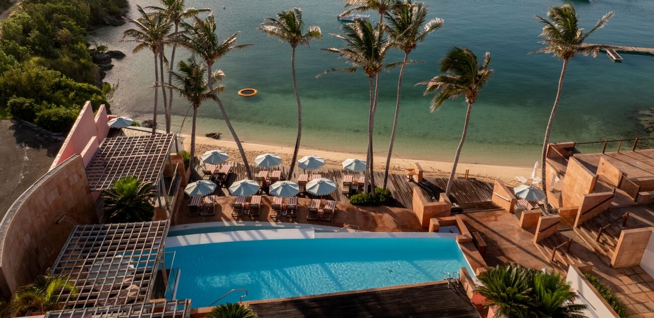 Colorful coastal resort with a pool, palm trees, beach, and boats on a calm bay.