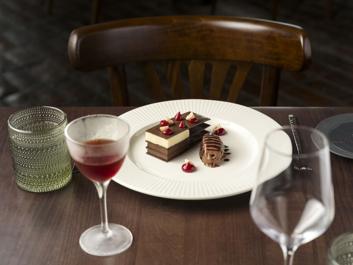 A chocolate dessert plate with layered cake, a chocolate square, and garnishes, plus a wine glass, a water glass, and a green patterned tumbler on a dining table.
