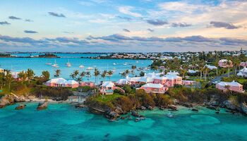 A tropical seaside village with pink-roofed cottages along turquoise waters and a calm harbor under a partly cloudy sky.