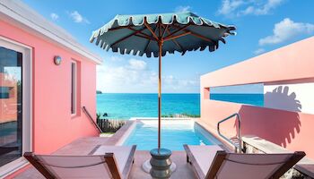 Beachfront resort with pink walls, turquoise sea, blue sky, and a central umbrella between two lounge chairs by a pool.