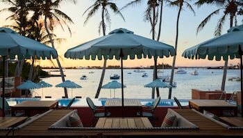 A tropical beach club at sunset with wooden loungers, turquoise umbrellas, and palm trees overlooking calm water and boats, dreamy and relaxing.
