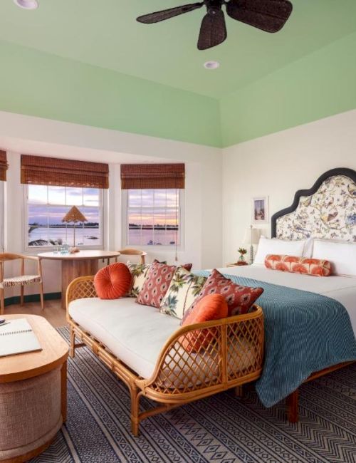 A cozy, well-decorated bedroom with a large bed, rattan furniture, and a scenic view from the bay windows overlooking the sea, ending the sentence.
