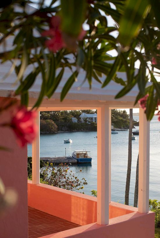 A view from a porch with pink flowers, overlooking a small dock and moored boats on a calm body of water, framed by tropical greenery.