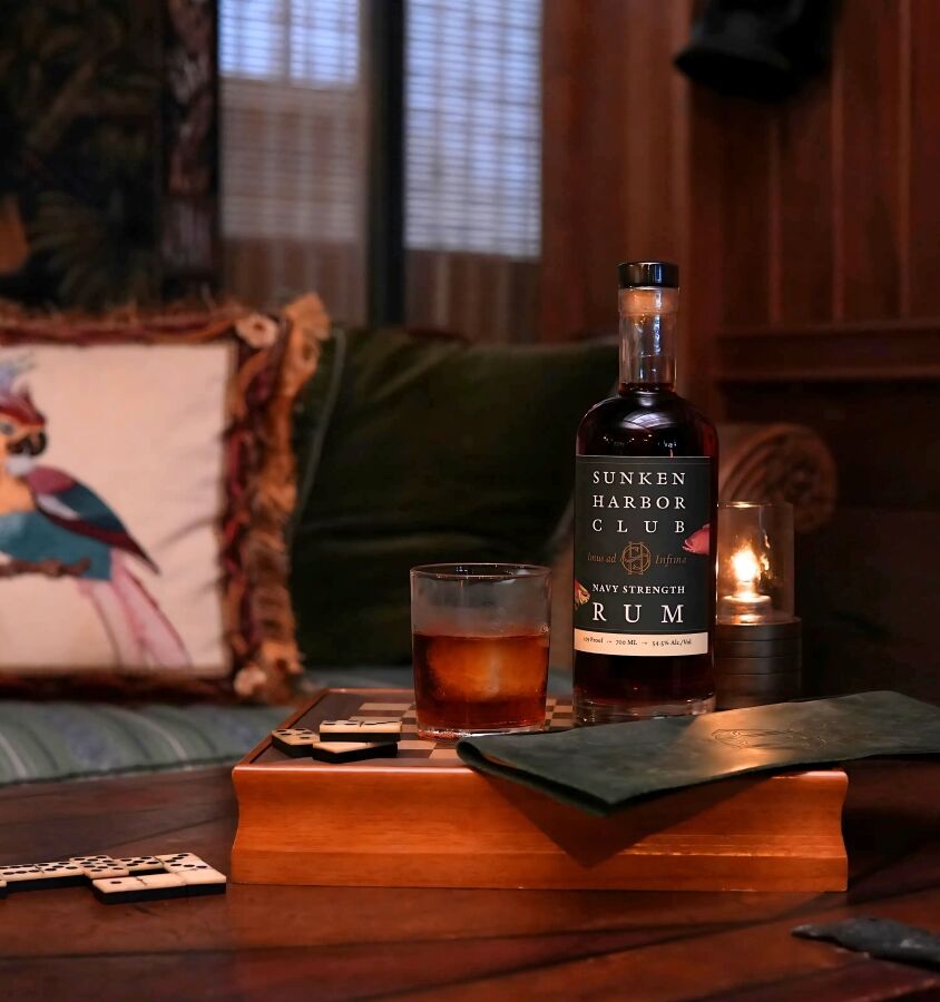 A cozy wooden table with a whiskey bottle, glass, and cards, a dark napkin, and a colorful pillow in the background, lamp-lit room.