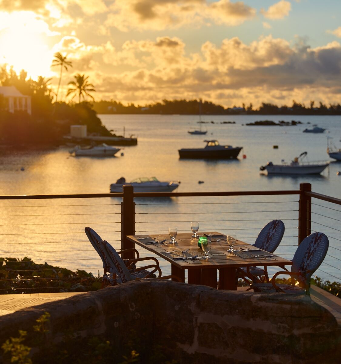 A sunset view over a calm harbor with boats, a terrace table and chairs, palm trees, and a warm, tropical glow.