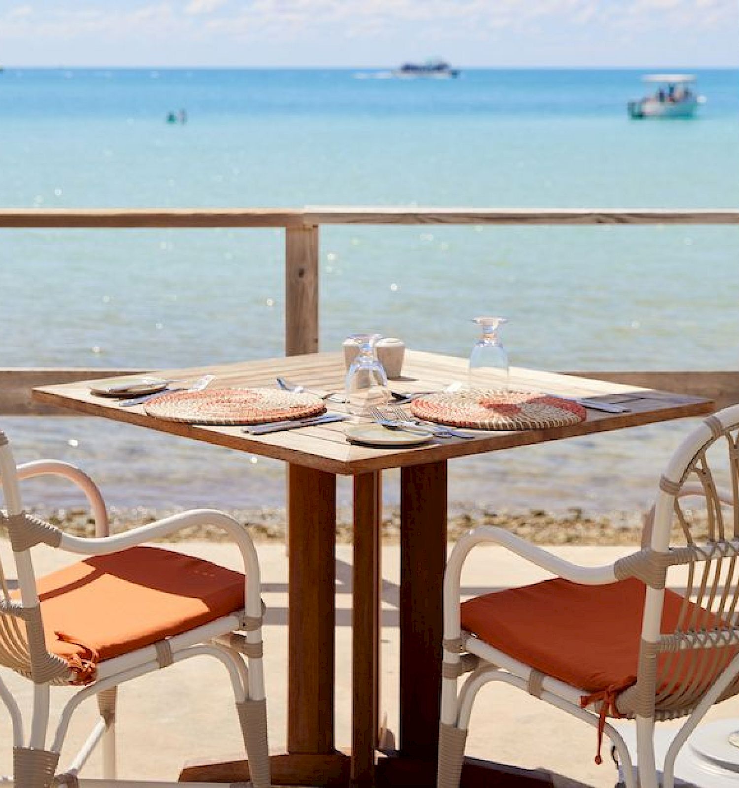 A beachside dining setup with two chairs and a table, featuring orange cushions and place settings, overlooking clear blue waters and boats.