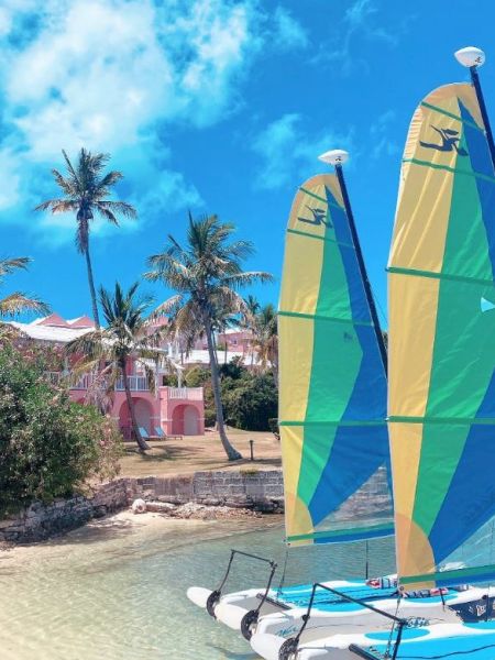 The image shows two colorful sailboats docked on a sandy beach with palm trees and colorful buildings in the background under a bright blue sky.