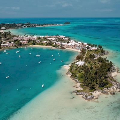 Aerial view of a coastal area with turquoise waters, boats docked, sandy beaches, palm trees, and buildings along the shoreline, under a blue sky.