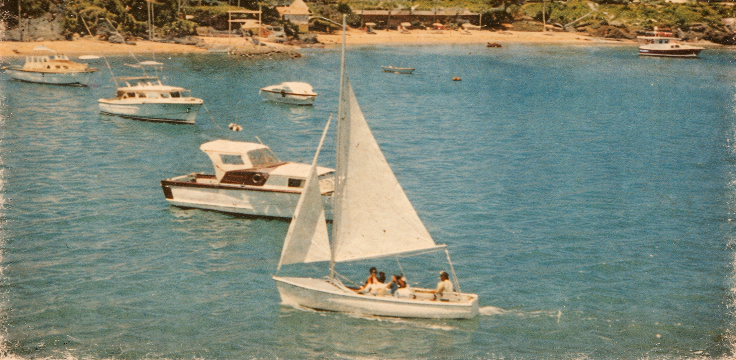 Sailing boats on calm turquoise water near a sunny beach with palm trees and resort buildings in the background, a peaceful seaside scene.