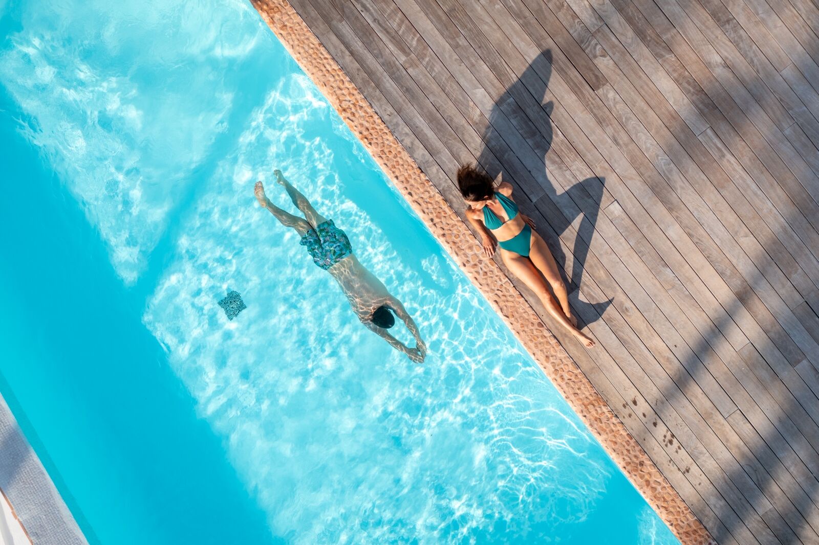 Two people relaxing by a bright blue pool: a woman sunbathing on the wooden deck and a person floating on their back in the water.