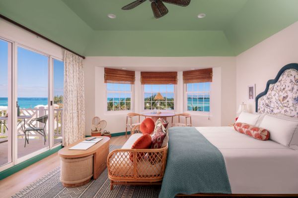 A cozy bedroom with tropical decor, featuring a large bed, a sitting area, and a balcony overlooking a stunning ocean view under a green ceiling.