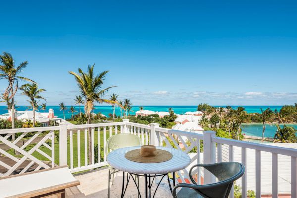 A balcony with a white table, two chairs, a hat, overlooking a tropical landscape with palm trees, and clear blue ocean under a bright sky.