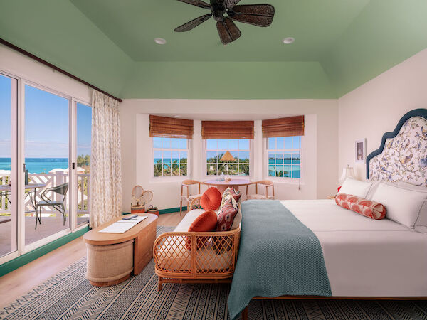 A cozy bedroom with tropical decor, featuring a large bed, a sitting area, and a balcony overlooking a stunning ocean view under a green ceiling.