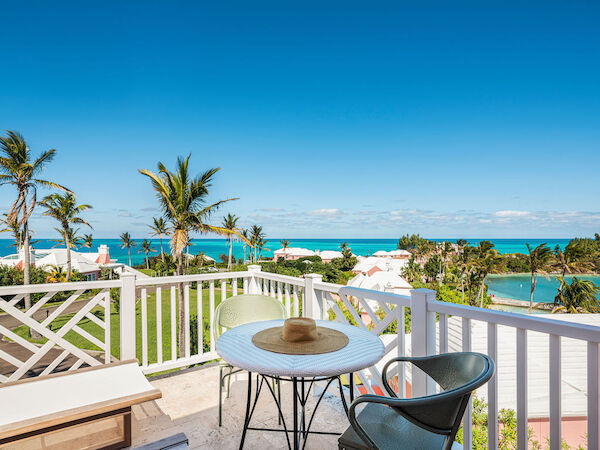 A balcony with a white table, two chairs, a hat, overlooking a tropical landscape with palm trees, and clear blue ocean under a bright sky.