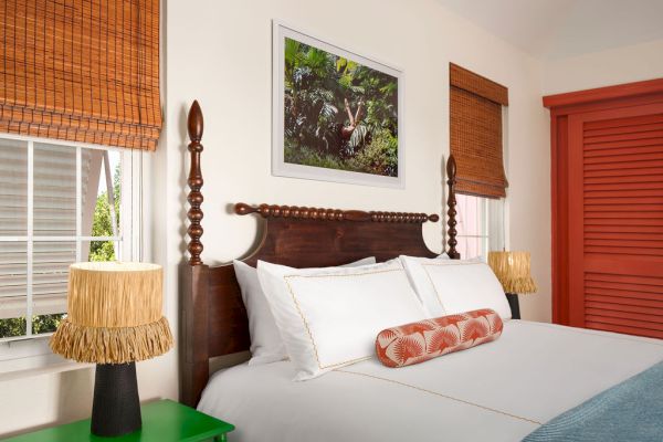 A cozy bedroom featuring a wooden bed with white linens, pillow, two lamps, a green nightstand, bamboo blinds, and artwork above the bed.