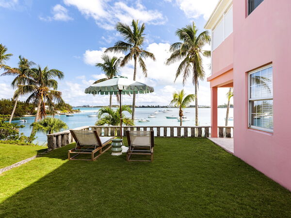 A seaside view with palm trees, lounge chairs under an umbrella, green grass, boats on the water, and a pink building on the right.