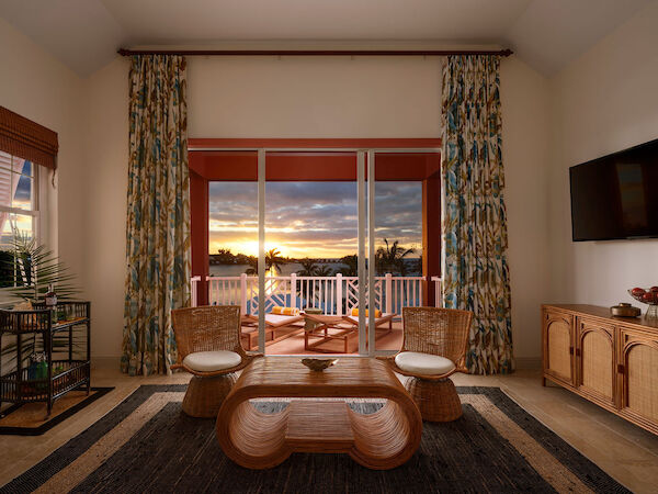 A cozy living room with wicker furniture, a wooden coffee table, a mounted TV, and large windows opening to a balcony and a sunset view, ending the sentence.