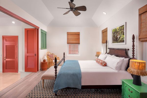 A cozy bedroom features a double bed with a wooden frame, colorful doors, wicker furniture, a green nightstand, and wicker blinds on the windows.