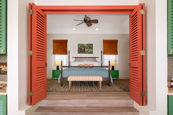 A cozy bedroom with a large bed, orange shutters, and green accents. The room is well-lit and features wooden blinds, a ceiling fan, and side tables.