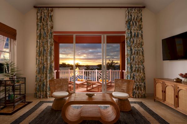 A cozy living room with wicker furniture, a wooden coffee table, a mounted TV, and large windows opening to a balcony and a sunset view, ending the sentence.