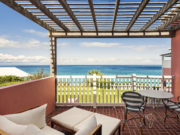 A cozy patio with a sofa, chairs, and a table, overlooking a grassy area and a beautiful ocean view through a wooden lattice.