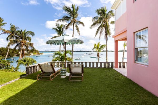 A seaside view with palm trees, lounge chairs under an umbrella, green grass, boats on the water, and a pink building on the right.