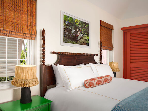 A cozy bedroom features a wooden bed with white linens, a colorful bolster pillow, bamboo shades, two lamps, green nightstand, and tropical wall art.