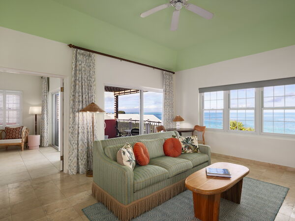 A living room with a green ceiling, patterned curtains, a green couch with orange pillows, a coffee table, and ocean view through the windows.