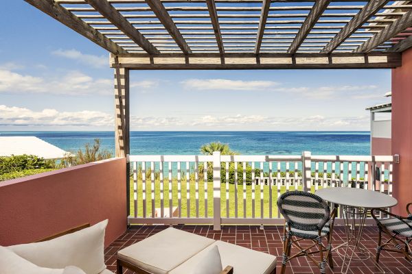 A patio with wooden furniture overlooks a grassy area, picket fence, and expansive ocean view. The sky is partly cloudy, providing a serene setting.