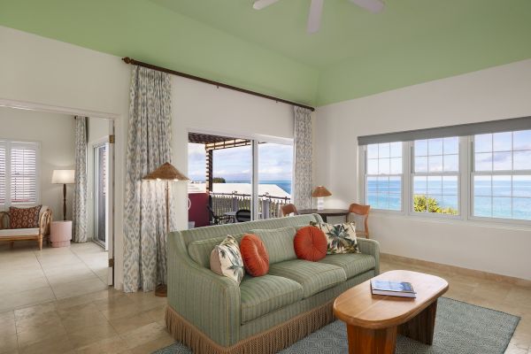 A living room with a green ceiling, patterned curtains, a green couch with orange pillows, a coffee table, and ocean view through the windows.