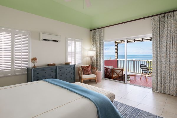 A cozy bedroom featuring a large bed, a chest of drawers, a chair, a lamp, and an open balcony with a view of the ocean.