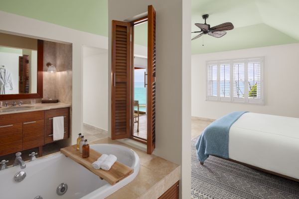 A stylish bathroom with a large bathtub and adjacent bedroom, featuring wooden decor and light airy colors, overlooking a sea view through windows.