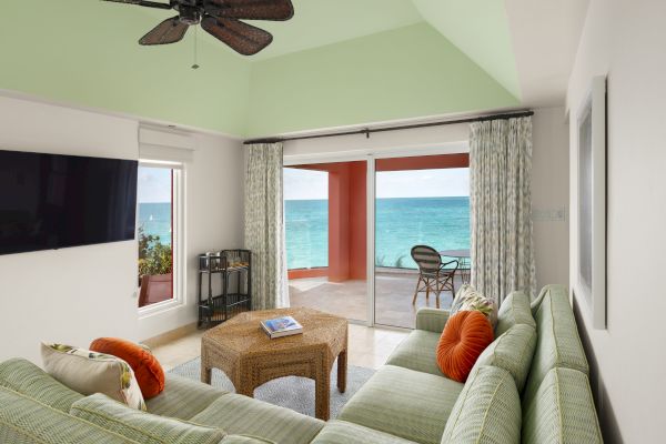 A cozy living room with green striped furniture, a wicker coffee table, ceiling fan, TV, and a scenic ocean view through sliding glass doors.