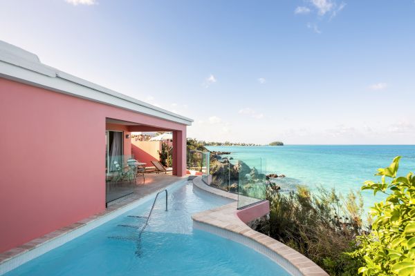 A picturesque oceanfront villa with a private pool and pink exterior, overlooking a clear blue sea under a sky with scattered clouds.