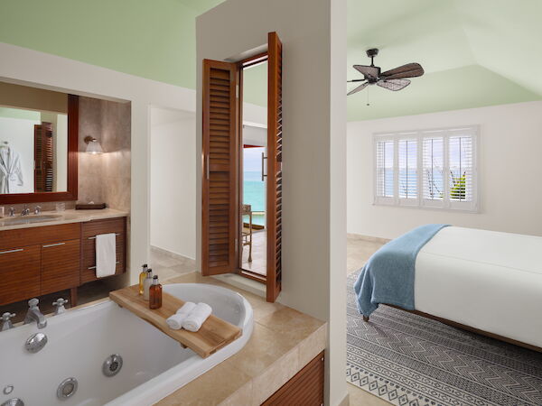 A stylish bathroom with a large bathtub and adjacent bedroom, featuring wooden decor and light airy colors, overlooking a sea view through windows.