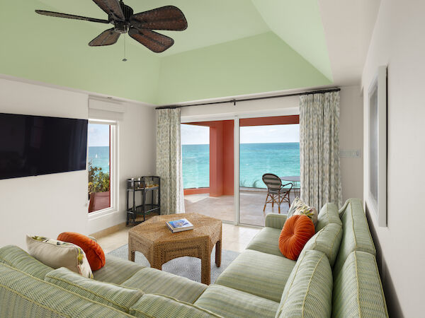 A cozy living room with green striped furniture, a wicker coffee table, ceiling fan, TV, and a scenic ocean view through sliding glass doors.