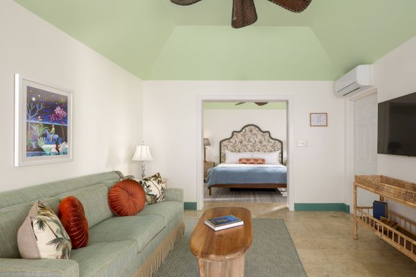 The image shows a living room with a green couch, coffee table, TV, and a view of a bedroom in the background with a bed and nightstand.