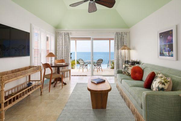 A cozy living room with green accents features a sofa, coffee table, desk, TV, and opens to an ocean-view balcony with chairs and a table.
