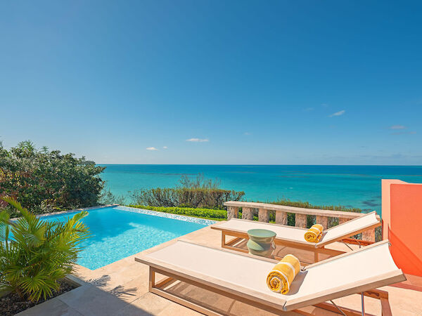 An ocean view from a poolside deck with two lounge chairs, towels, plants, and a clear blue sky in the background, creating a serene atmosphere.