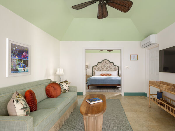 The image shows a living room with a green couch, coffee table, TV, and a view of a bedroom in the background with a bed and nightstand.