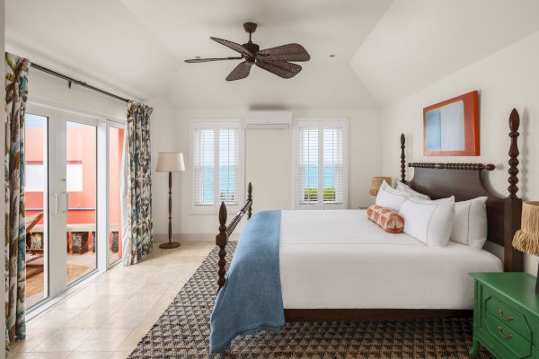 A spacious bedroom features a large bed, a ceiling fan, a green nightstand, and glass doors leading to a balcony with an ocean view.