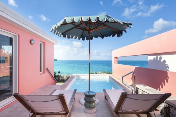 Image shows two lounge chairs under a large umbrella next to a pool, with an ocean view, flanked by pink walls under a clear blue sky.