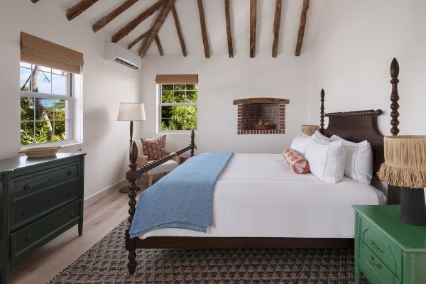 A cozy bedroom with wooden beams, a large bed, green dresser, nightstand, lamp, and windows letting in natural light, adorned with décor.