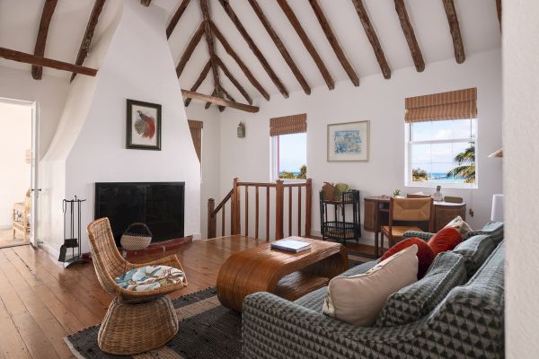 A cozy living room with rustic beams, a fireplace, a sofa, a wicker chair, a coffee table, and artwork, overlooks a scenic view through two windows.