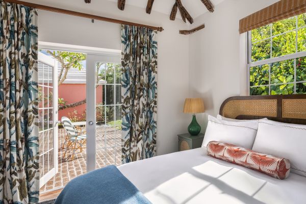 A cozy bedroom with exposed beams, a comfy bed, patterned curtains, a lamp, and French doors opening to a sunny patio with outdoor seating, ends the sentence.