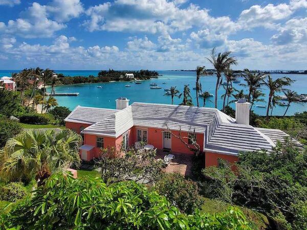 A house surrounded by lush greenery sits near a blue body of water with a picturesque view of the sky and clouds.