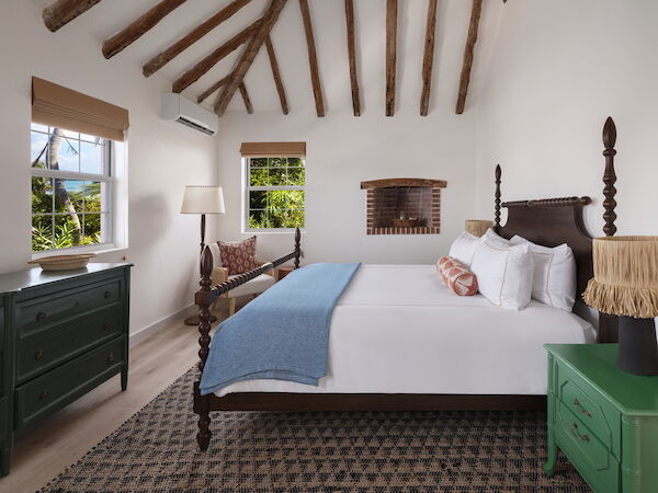 A cozy bedroom with wooden beams, a large bed, green dresser, nightstand, lamp, and windows letting in natural light, adorned with décor.