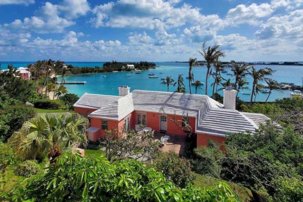 A house surrounded by lush greenery sits near a blue body of water with a picturesque view of the sky and clouds.