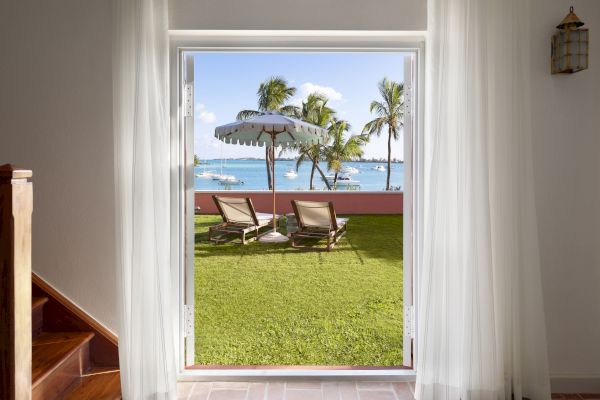 A doorway opens to a view of a grassy yard with lounge chairs under an umbrella, overlooking a beach with palm trees and boats on the water.